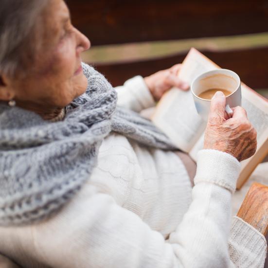 Ældre kvinde sidder og læser bog og drikker kaffe.