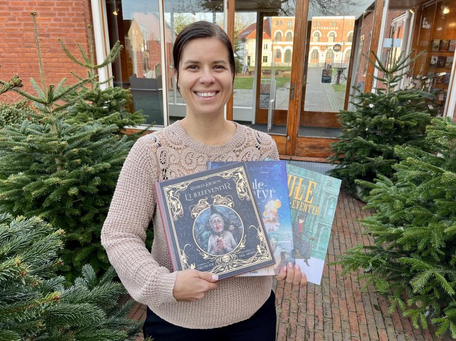 Marianne står omgivet af grantræer bag Nyborg Bibliotek, hvor hun holder de tre udgaver af "Et Juleeventyr", som hun anbefaler.